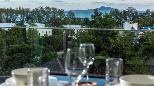 A dining table with plates, glasses, and cutlery set against a backdrop of a lush green landscape and a view of the sea. - Best Sea View 500m 2 Beach Modern C1 (Rawai)