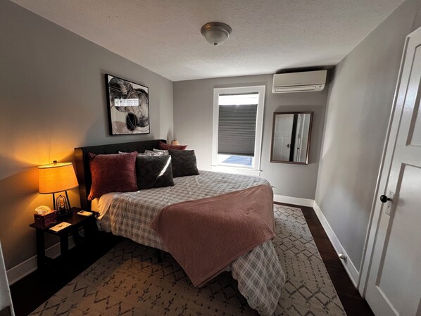 A bedroom with a bed, bedside tables, a lamp, a window with blinds, a ceiling light, and a wall-mounted air conditioner.