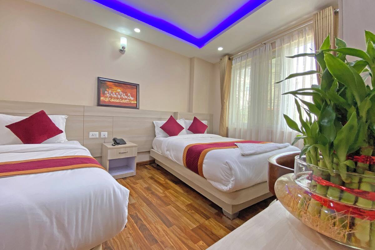 A hotel room with two beds, a nightstand, a phone, a painting, and a large plant.