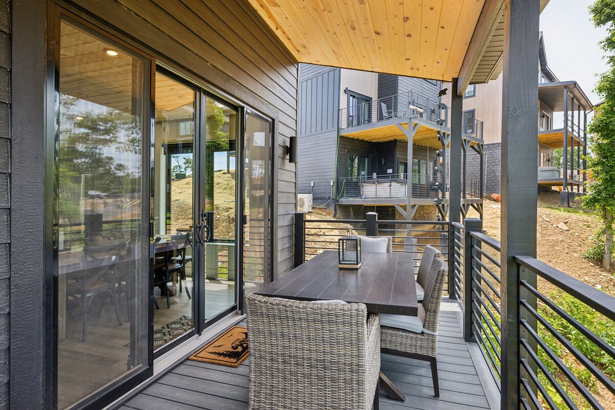 A modern outdoor deck with a dark wooden table and wicker chairs, connected to a building with large glass doors and a wooden ceiling.