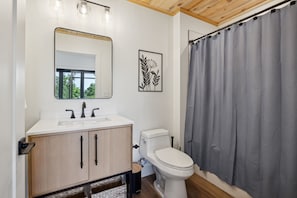 Un baño moderno con un lavabo blanco, un mueble de madera y un inodoro. Hay un espejo sobre el lavabo y una cortina de ducha a la derecha.