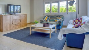 A modern living room with a sofa, a coffee table, a TV on a cabinet, and a window with a view of greenery.
