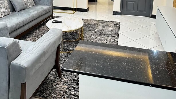 Ein modernes Wohnzimmer mit einem grauen Sofa, einem runden Couchtisch und einem schwarzen Hocker. Ein kleiner weißer Konsolentisch mit einer Blumenvase steht darin. Der Raum hat einen gefliesten Boden und eine graue Tür im Hintergrund.