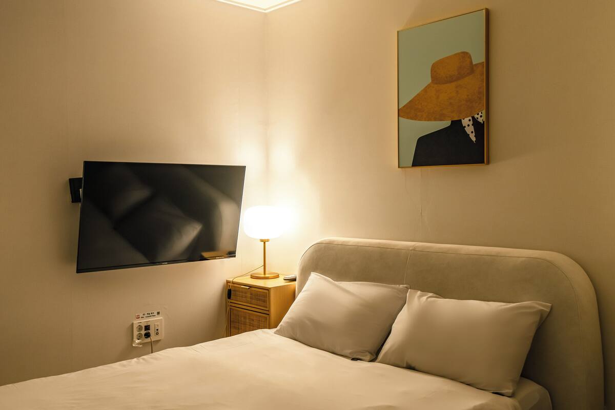 Una camera d'albergo con un letto, una televisione a parete, un comodino con una lampada e un quadro raffigurante un cappello.