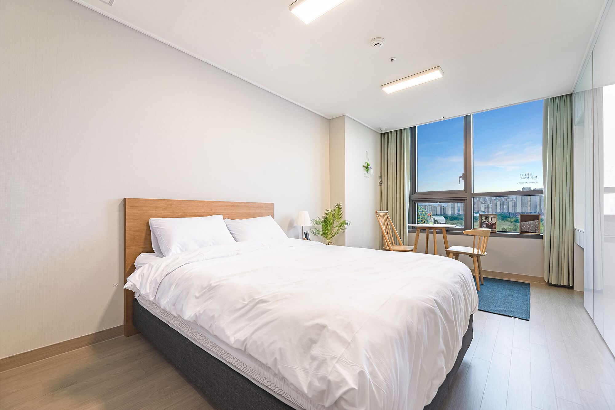 A modern hotel room with a large bed, a small table, and a view of the city through the window.