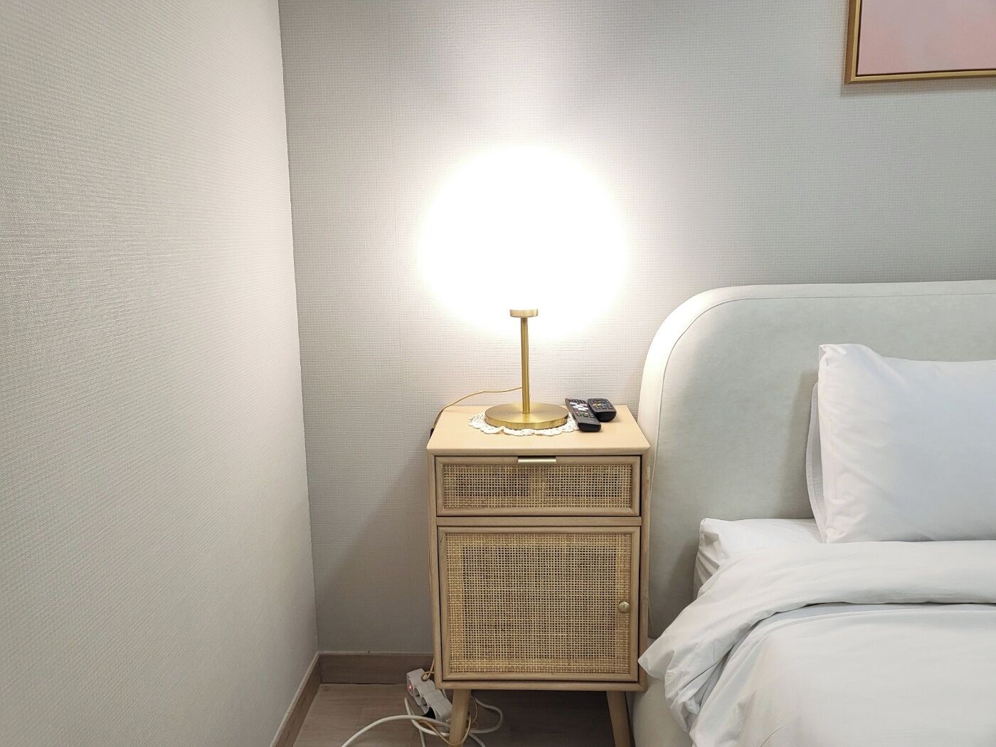 A neatly made bed with white bedding, a bedside lamp, and a small bedside table with a lamp and two remote controls.