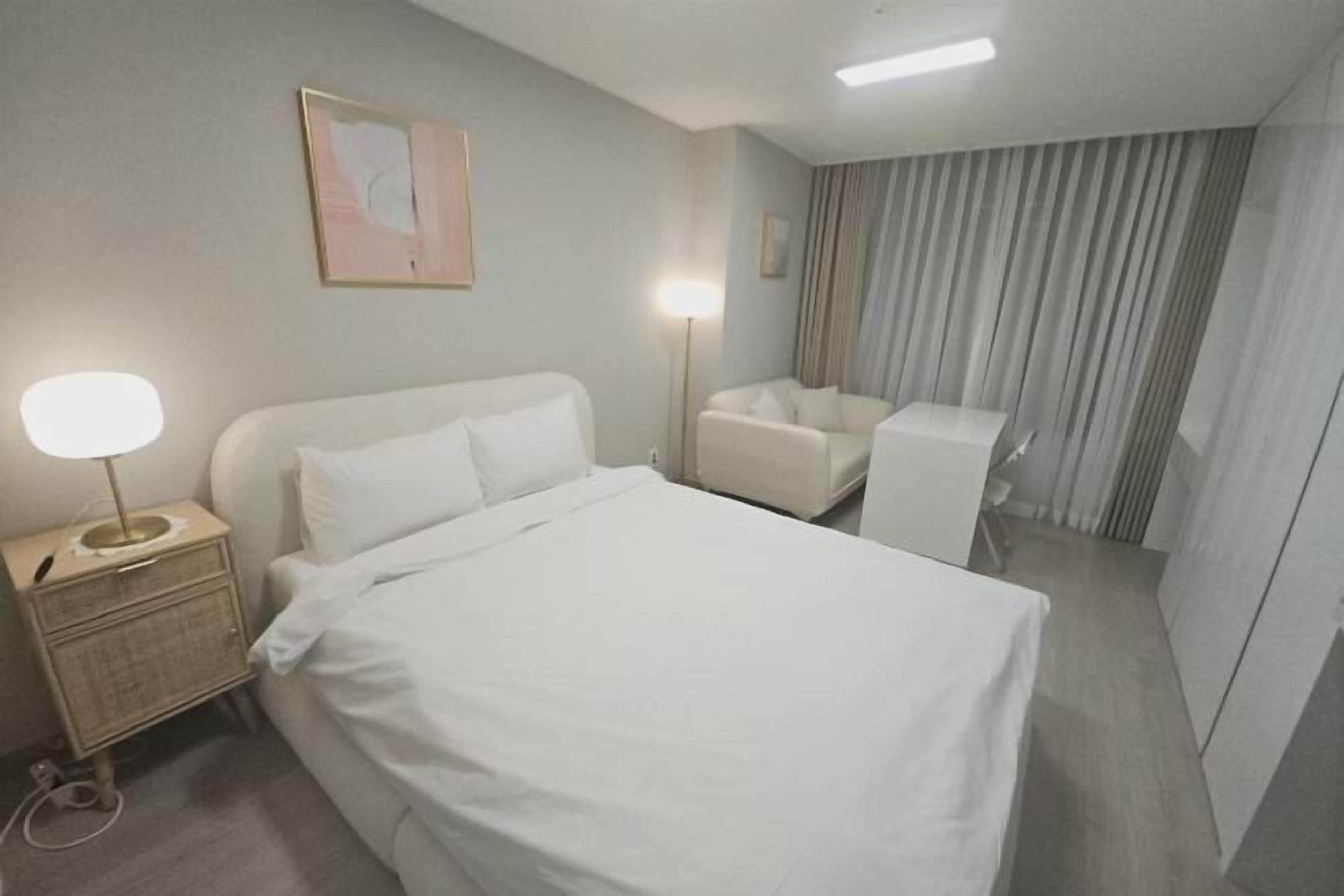 A hotel room with a bed, a bedside table with a lamp, a chair, and a large window with curtains.