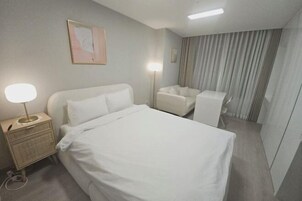 A hotel room with a bed, a bedside table with a lamp, a chair, and a large window with curtains.
