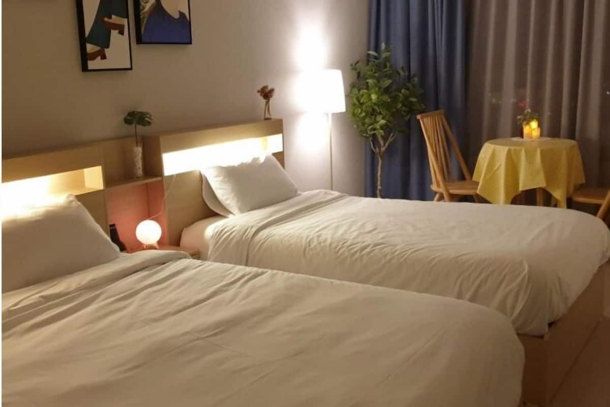 Camera d'albergo con due letti singoli, un comodino con una lampada, un piccolo tavolino rotondo con tovaglia gialla e una pianta in vaso.