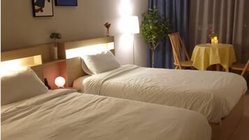 Une chambre d’hôtel avec deux lits simples, une table de chevet avec une lampe, une petite table ronde recouverte d’une nappe jaune et une plante en pot.