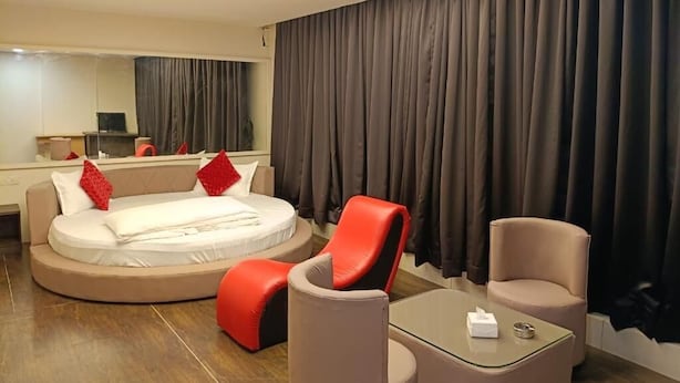 Ein modernes Hotelzimmer mit einem runden Bett, roten und beigen Stühlen, einem Glastisch und einem großen Fenster mit Vorhängen.