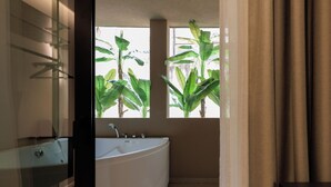 A modern bathroom with a large bathtub, a glass shower enclosure, and windows with tropical plants.