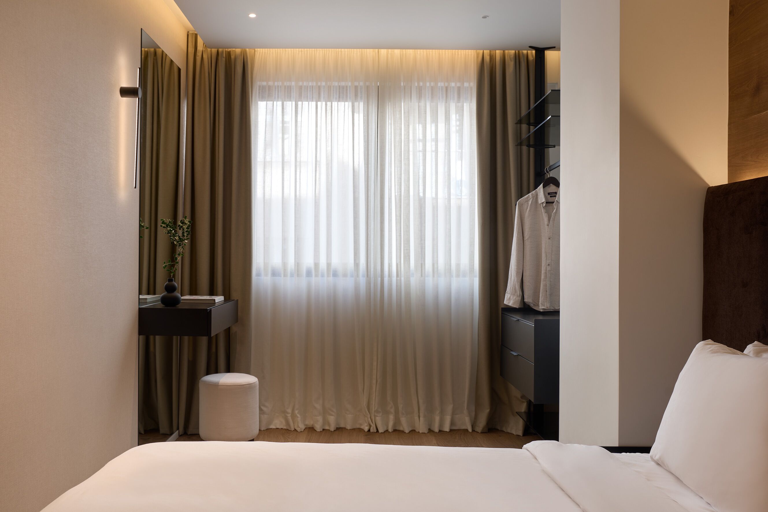 A hotel room with a bed, a bedside table, a mirror, a hanging coat rack, and a window with curtains.
