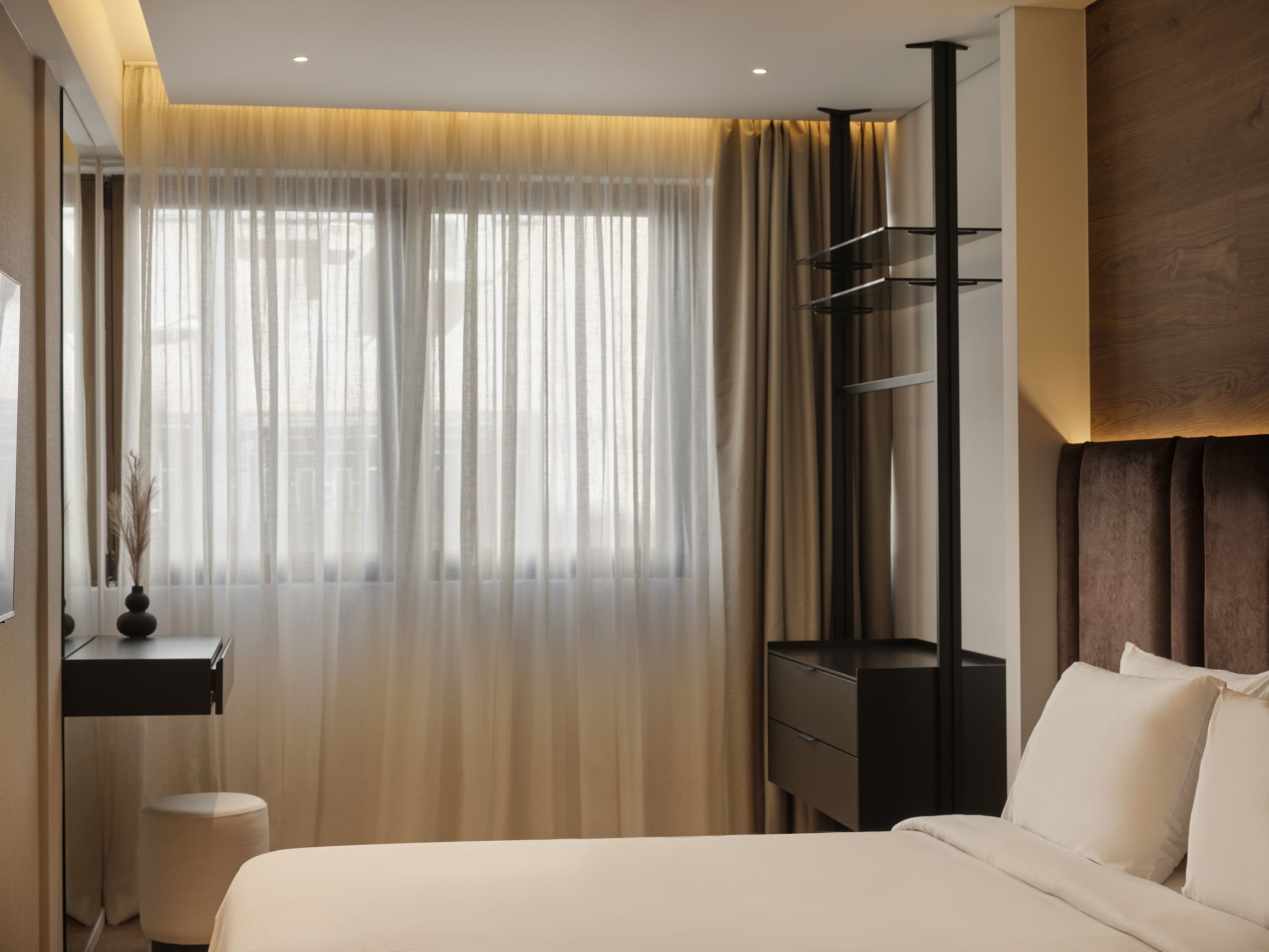 A modern hotel room with a bed, a desk, a chair, and a large window with sheer curtains.