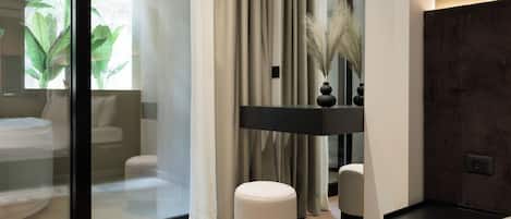 A modern bedroom with a glass partition, a round ottoman, a bedside table, a lamp, and a mirror.
