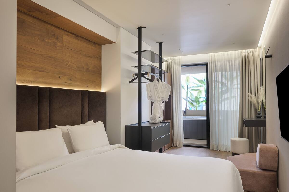 A modern bedroom with a large bed, a wooden headboard, a black dresser, a robe, a chair, and a TV.