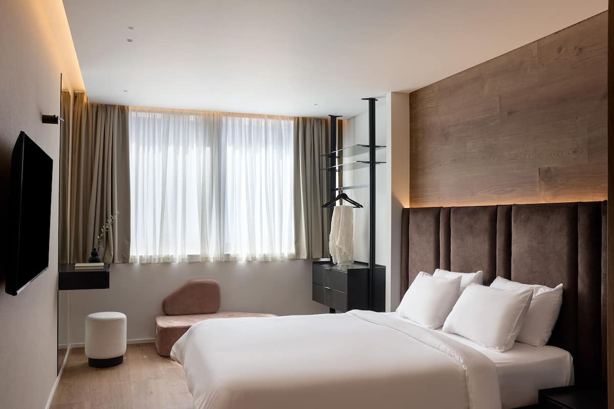 A modern hotel room with a large bed, a flat-screen TV mounted on the wall, a bedside table, a small round ottoman, a wooden headboard, and a window with sheer curtains.