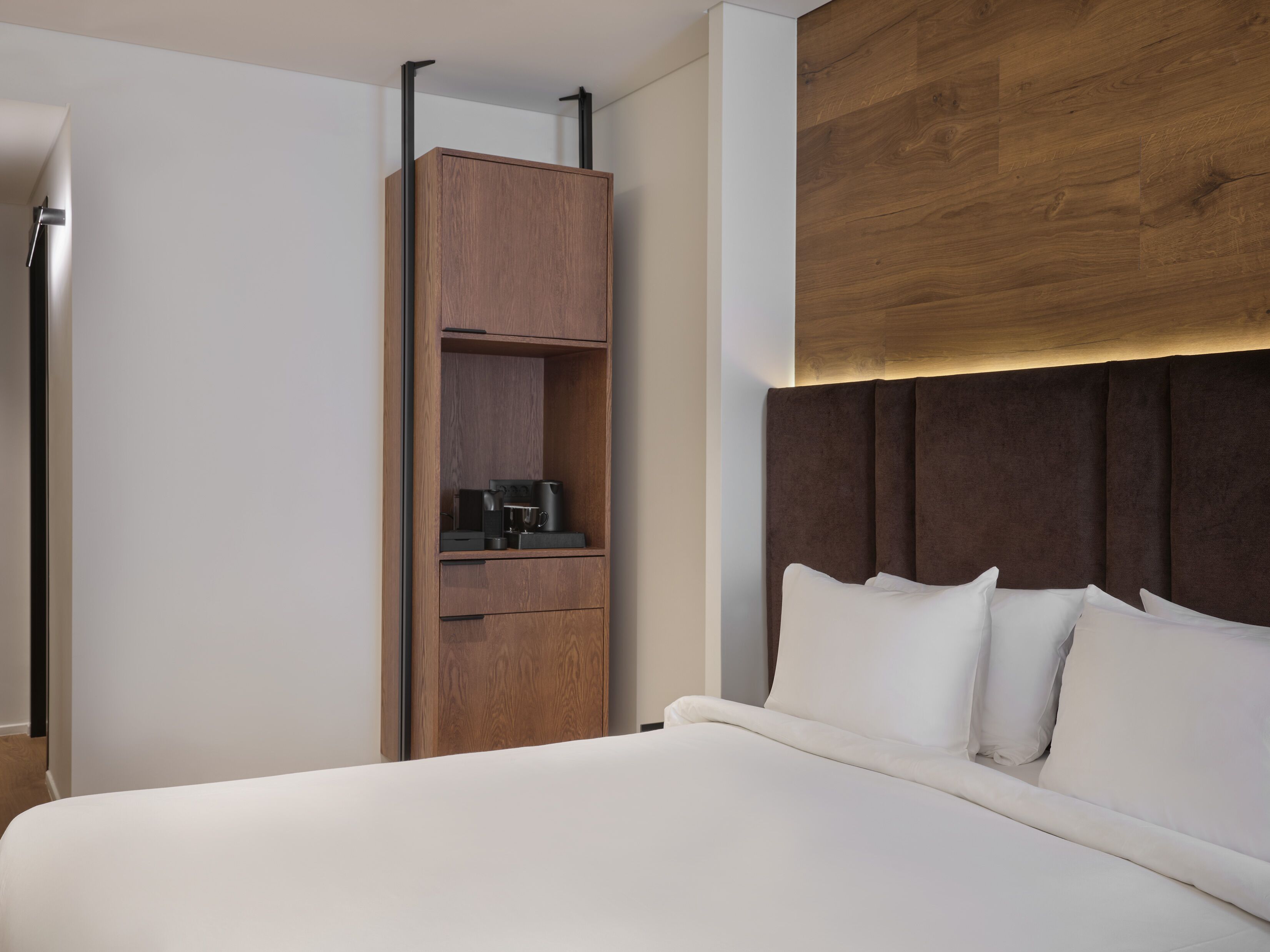 Una habitación de hotel moderna con una cama grande, un armario de madera y un cabecero con iluminación integrada.