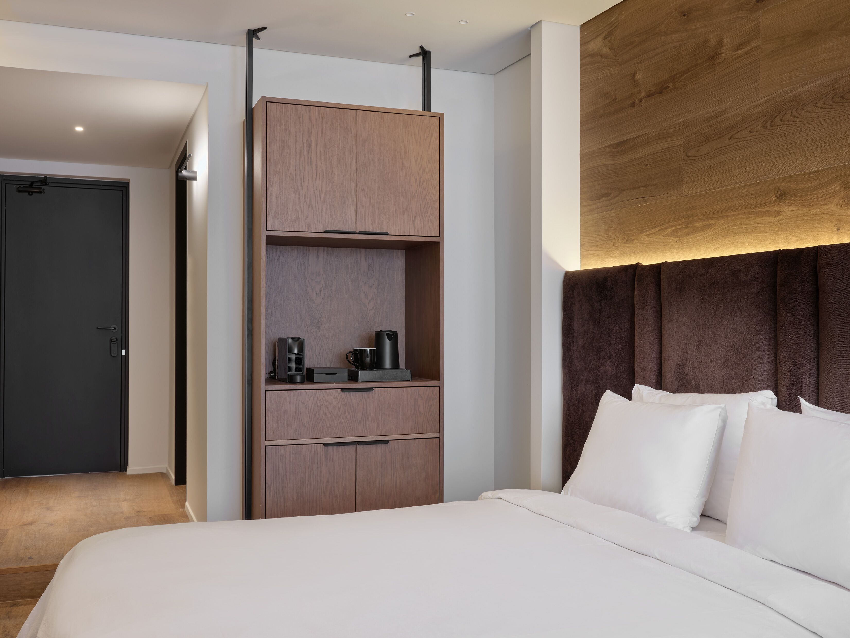 A hotel room with a bed, a wardrobe, a desk, and a dark door.