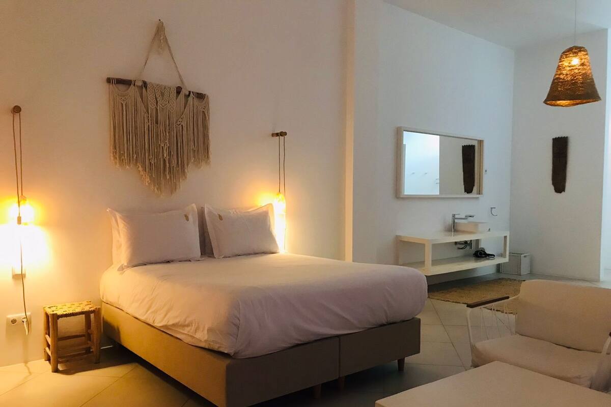 Un dormitorio con cama, lámparas de noche, mesita de noche, una silla, escritorio, espejo y un adorno colgante de macramé.