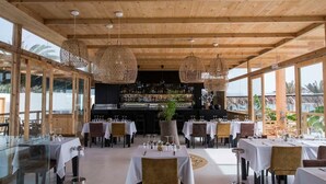 Un restaurant avec des poutres en bois, de grandes fenêtres et des lustres en osier suspendus au plafond. Les tables sont dressées avec des nappes blanches et des chaises.