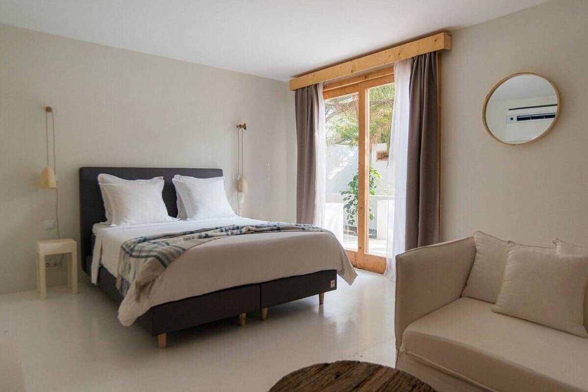 Un dormitorio con cama, mesita de noche, sofá, ventana con cortinas y un espejo redondo en la pared.