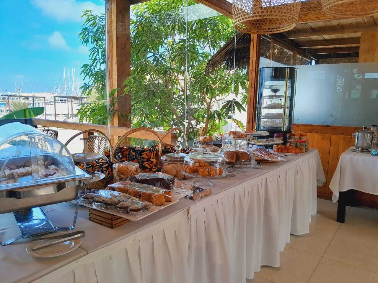 A buffet table with various food items and a grill, set in a covered outdoor area with a view of the sea and buildings in the background.