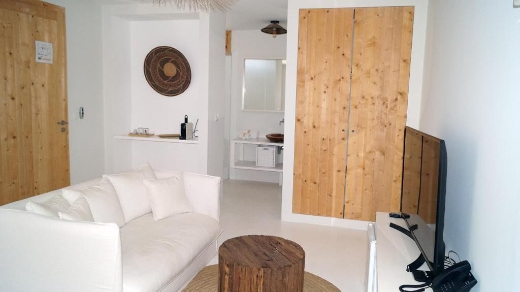 A modern living room with a white sofa, a wooden coffee table, and a TV. There is a wooden wardrobe and a mirror above the sink.