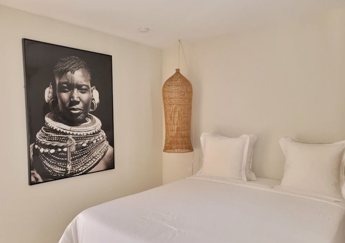 A bedroom with a large bed, white bedding, a wall-mounted artwork, and a woven light fixture.