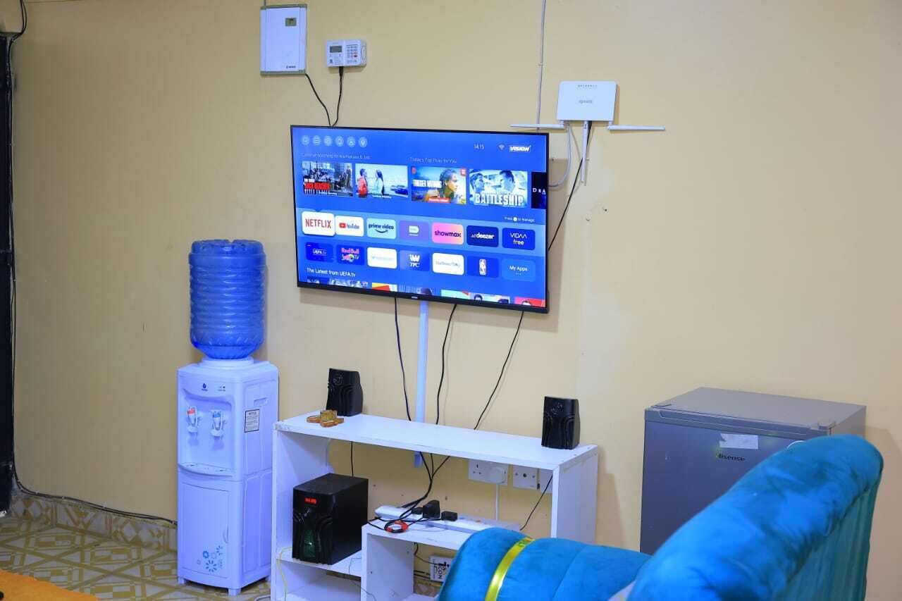 Una habitación con una televisión montada que muestra diversos servicios de streaming, un dispensador de agua, un escritorio blanco con una computadora y parlantes, y un refrigerador pequeño.