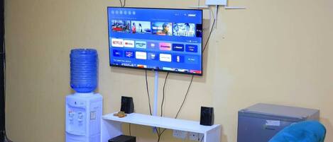 Una habitación con una televisión montada que muestra diversos servicios de streaming, un dispensador de agua, un escritorio blanco con una computadora y parlantes, y un refrigerador pequeño.