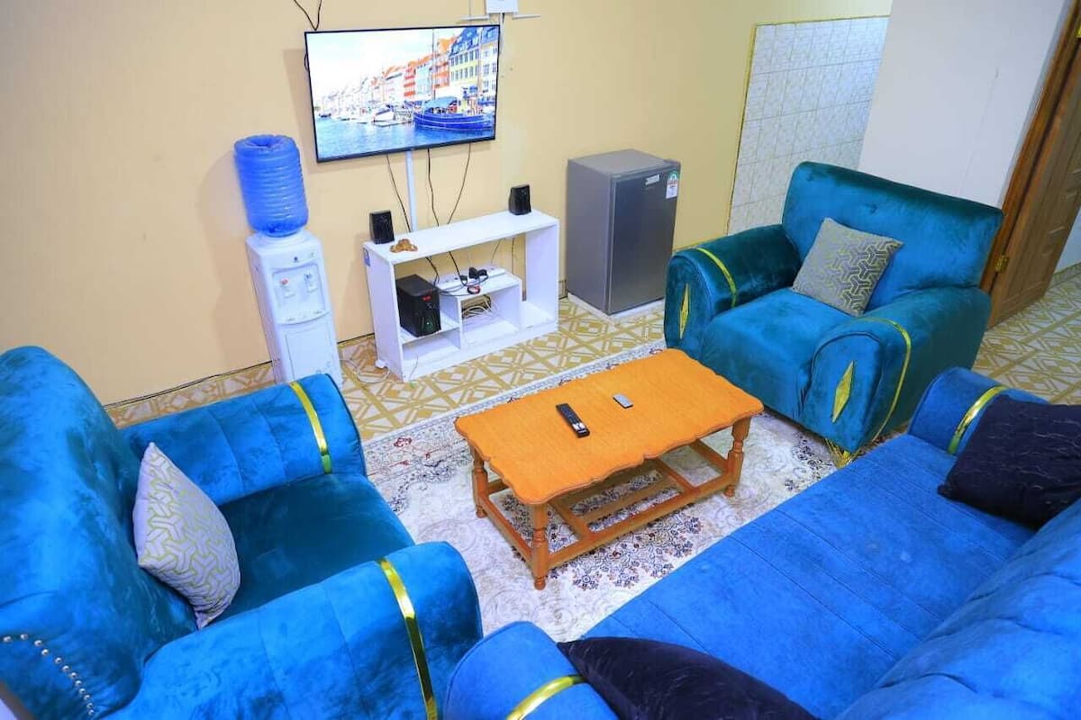 A living room with blue sofas, a wooden coffee table, a TV, a water dispenser, and a mini-fridge.