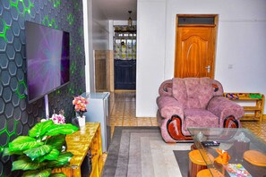 A living room with a large flat-screen TV, a pink armchair, a glass coffee table, and a wooden side table with a plant. - Lux Suites RFH Apartments Ruiru (Ruiru)