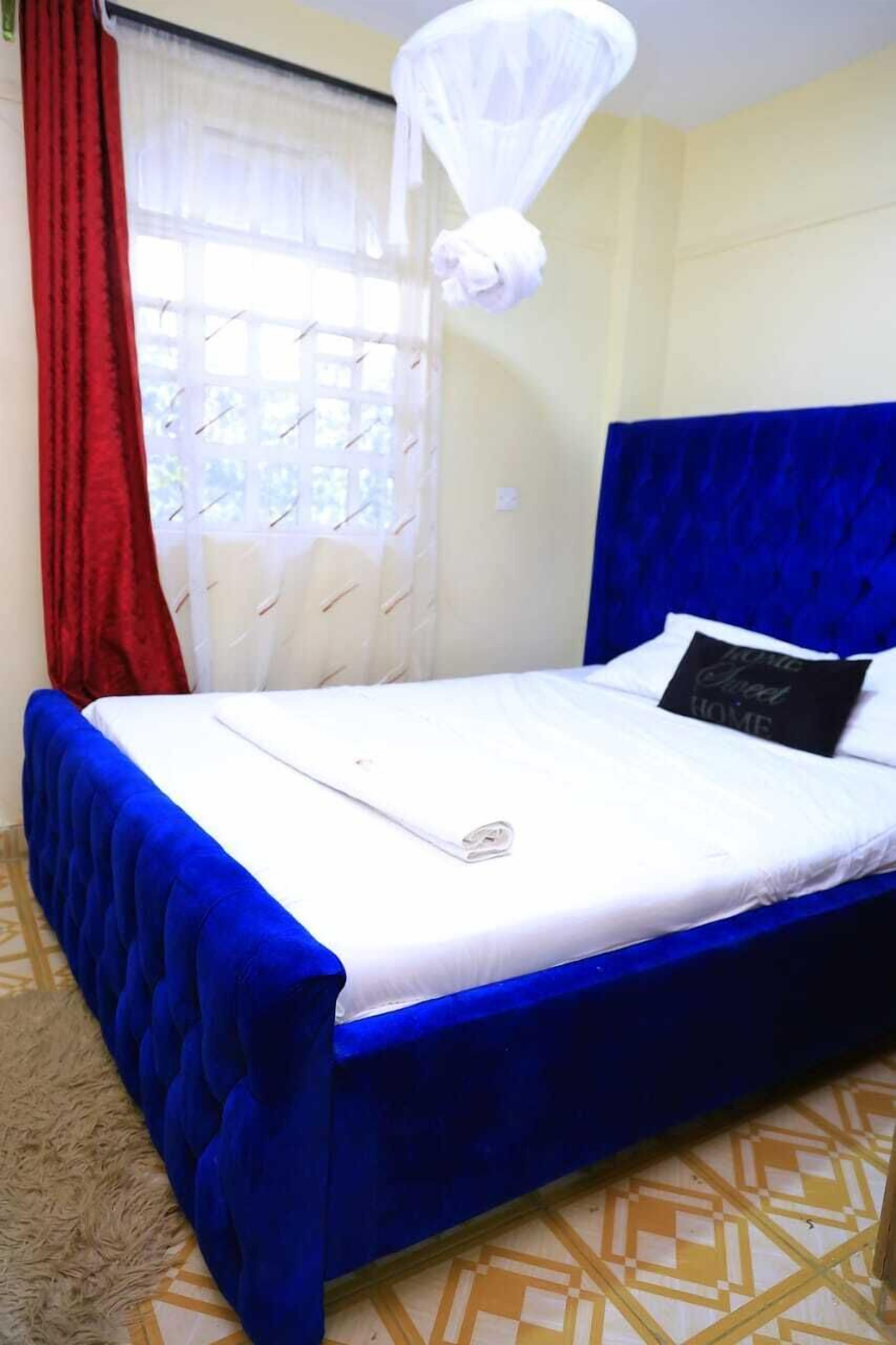 Een bed met een blauw hoofdeinde, witte beddengoed, een rood gordijn en een wit muggengaas.