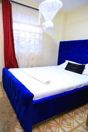 A bed with a blue headboard and white bedding, a red curtain, and a white mosquito net. - Lux Suites RFH Apartments Ruiru (Ruiru)