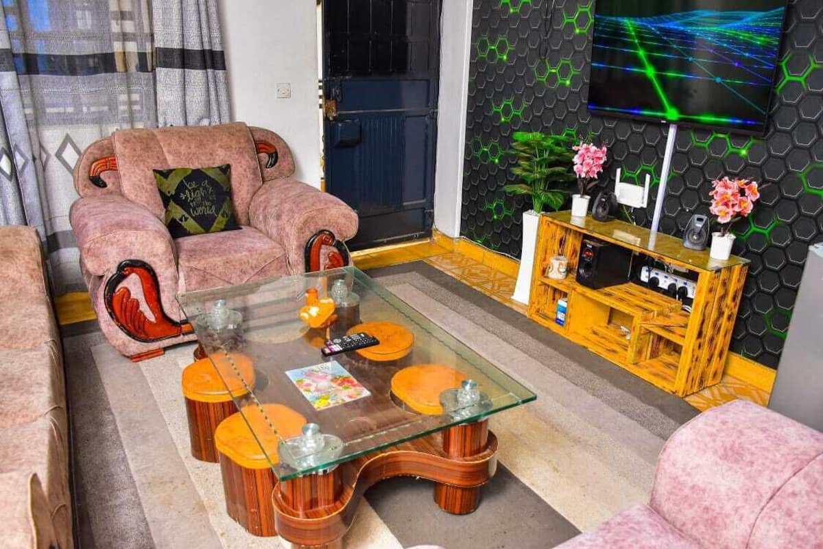 A living room with a glass coffee table, a pink sofa, a wooden entertainment unit, and a TV on a hexagonal wall.