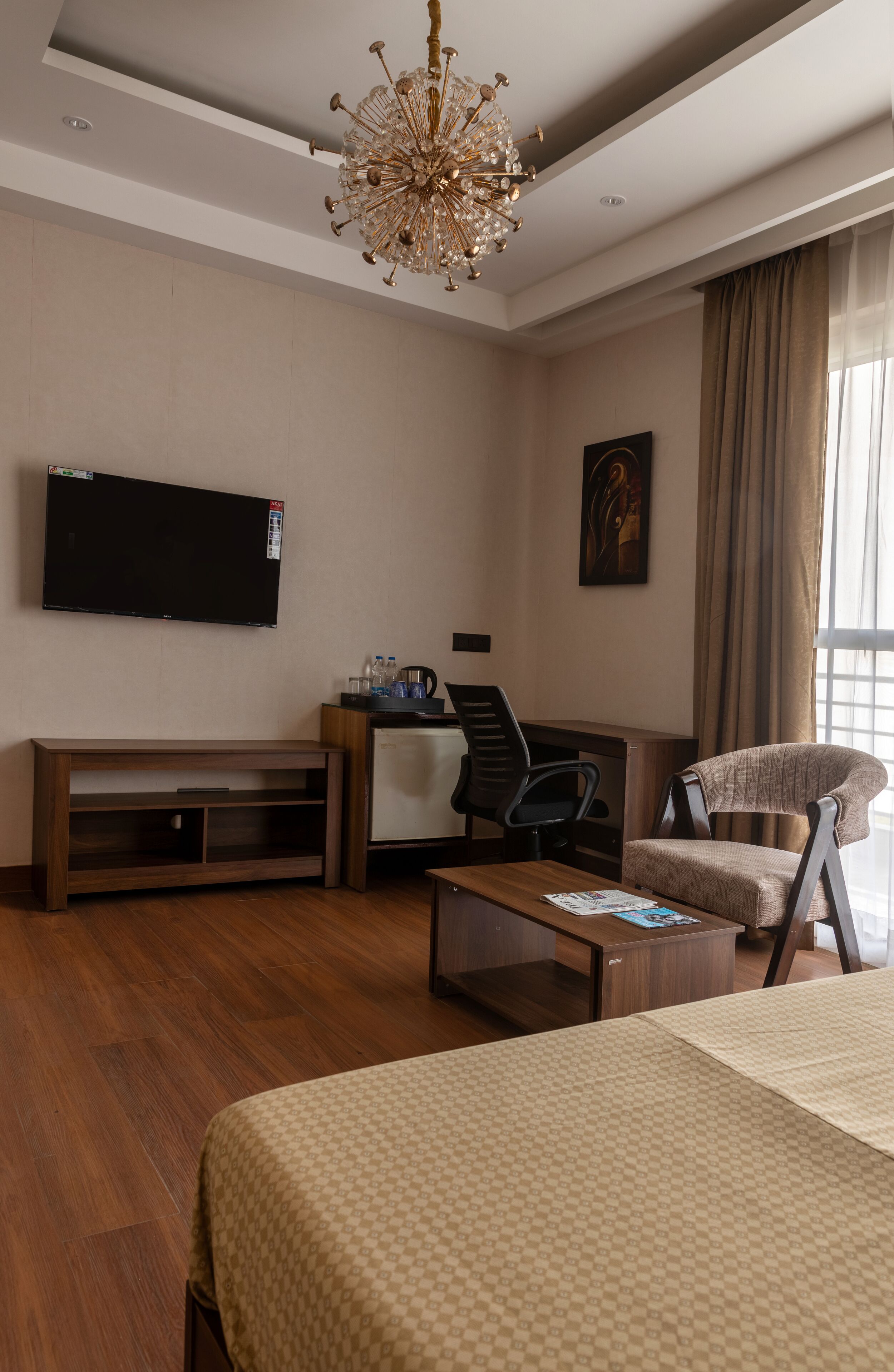 A modern hotel room with a large bed, a wooden coffee table, a TV mounted on the wall, a desk with a chair, and a window with curtains.