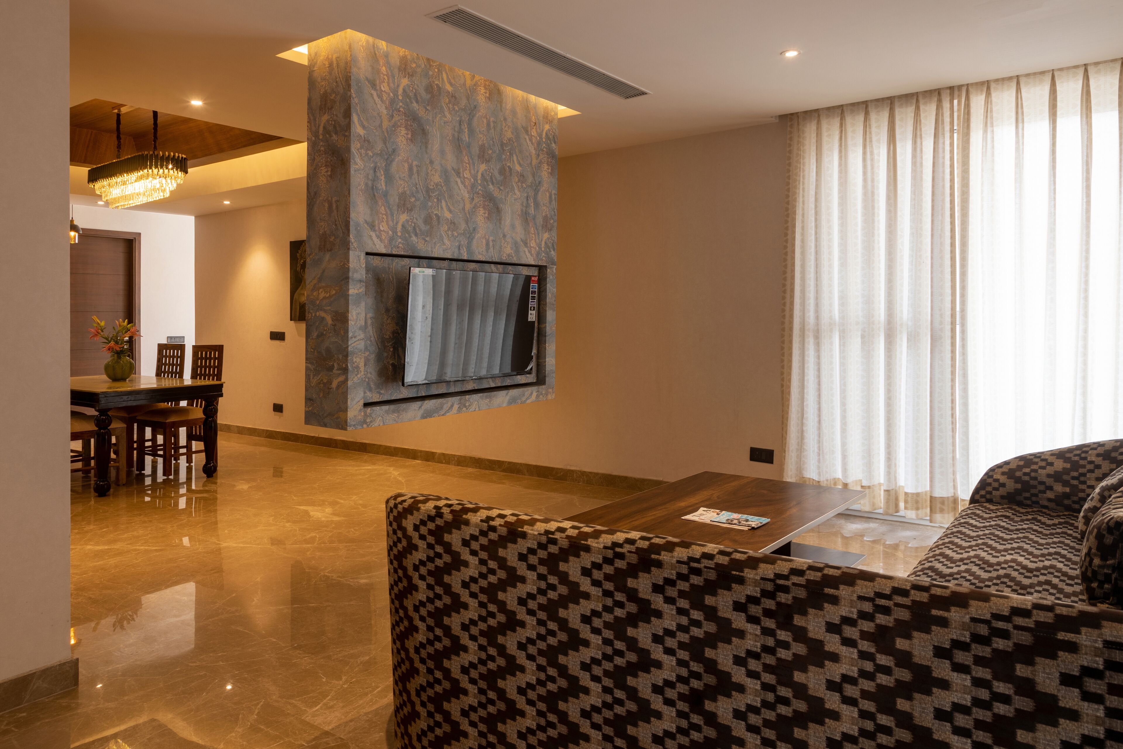 Un soggiorno moderno con una TV a schermo piatto montata su una parete strutturata, un divano con motivo decorativo, una zona pranzo con tavolo e sedie in legno e un'ampia finestra con tende leggere.
