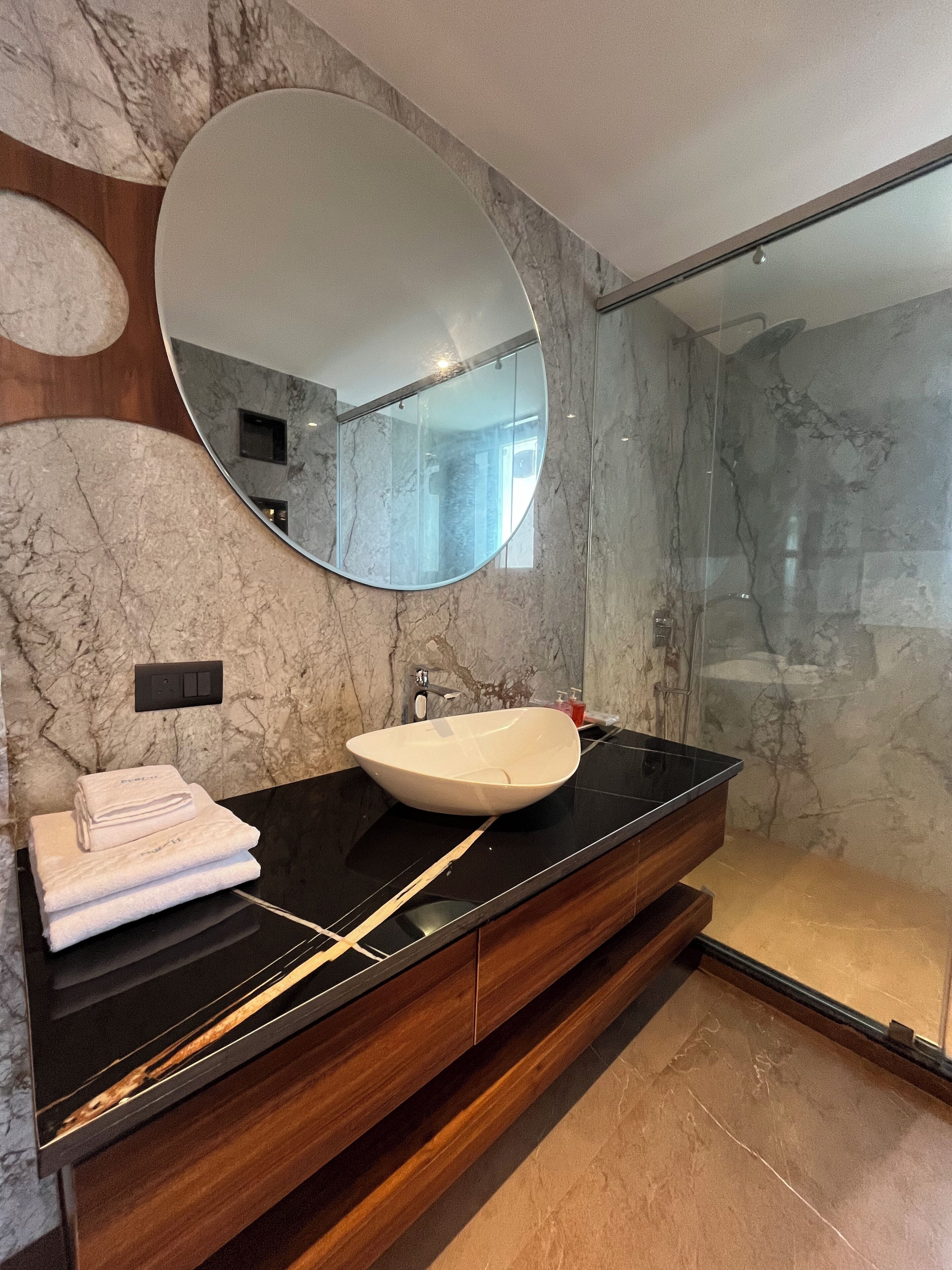 Ein modernes Badezimmer mit Marmorwand, einem großen Spiegel, einem weißen Waschbecken und einer Duschkabine aus Glas.