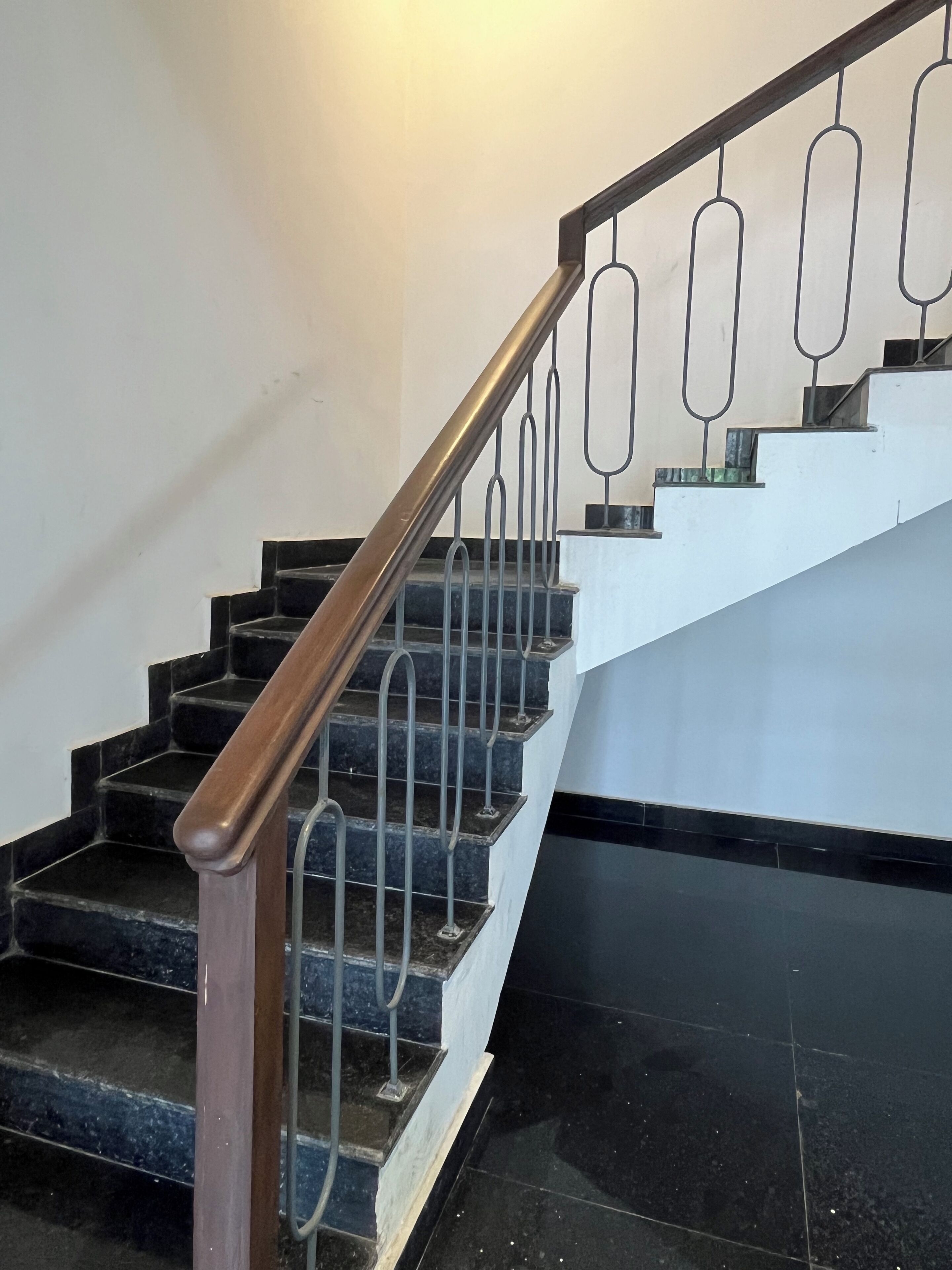 Una scala con ringhiera in legno e balaustre metalliche, che porta a un pianerottolo con parete bianca e pavimento scuro.