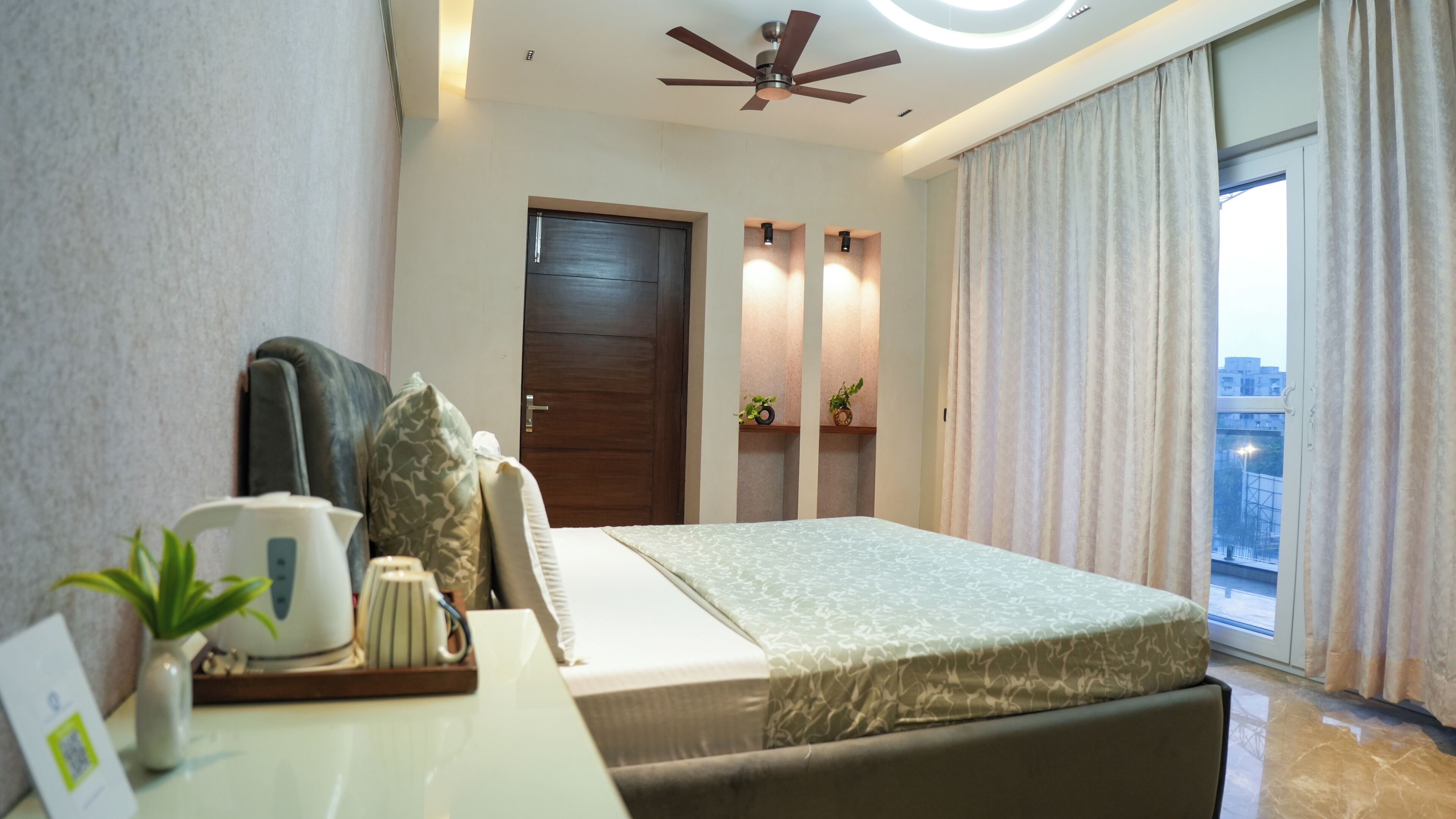 A hotel room with a bed, a desk with a plant, a ceiling fan, and a view of the outside.