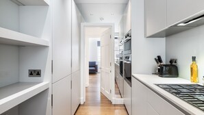 A narrow kitchen with white cabinetry, a wooden floor, and a view into a living area. - Stylish 2BR 2BA by the Heart of Soho (London)