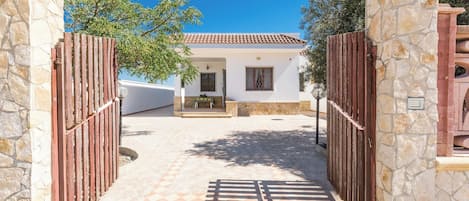 A gated entrance with stone walls and a tiled pathway leading to a white building with a tiled roof.