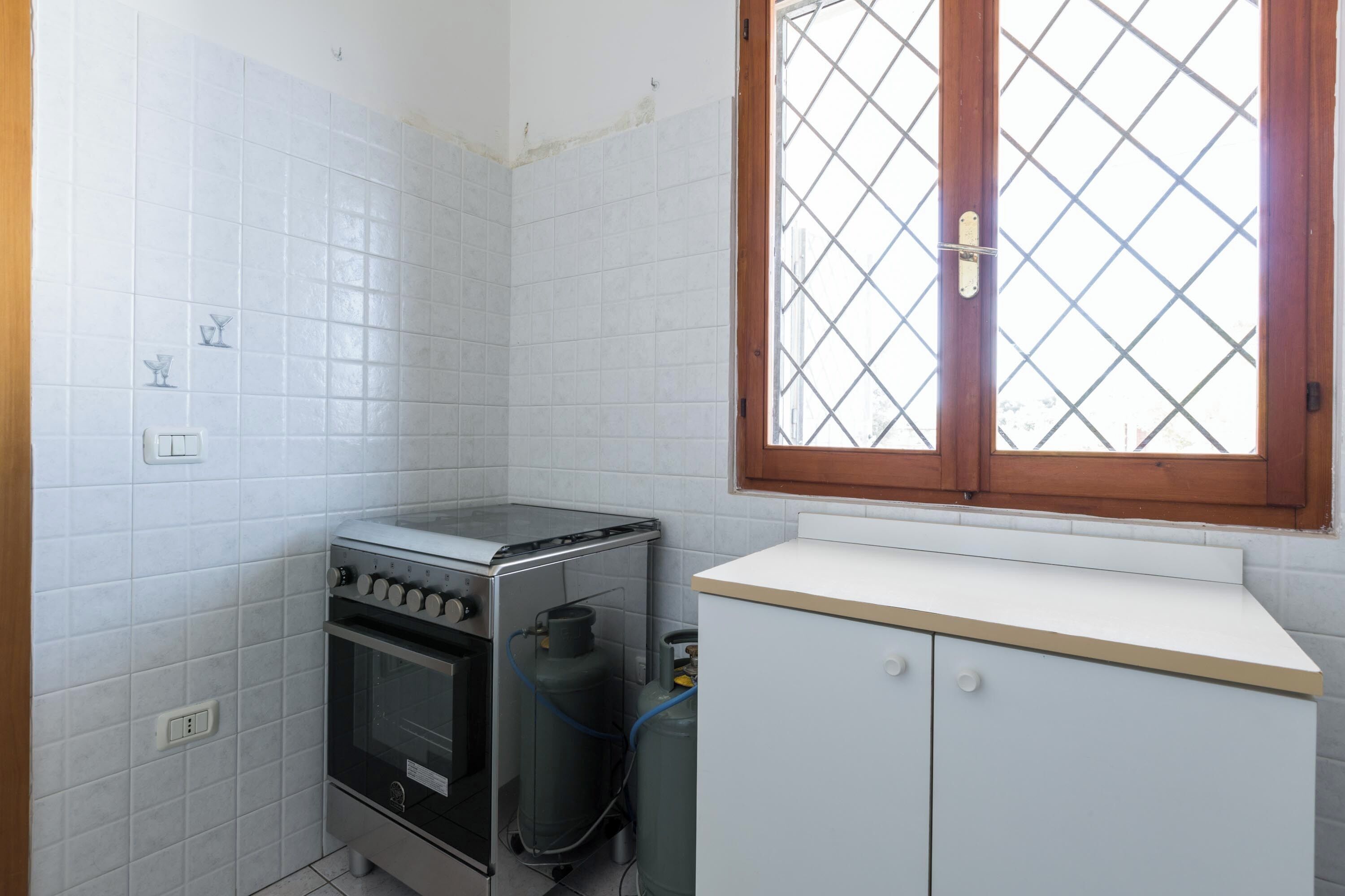 A compact kitchen with white tiles, a gas stove, a small oven, and a white cabinet below a window with a wooden frame.