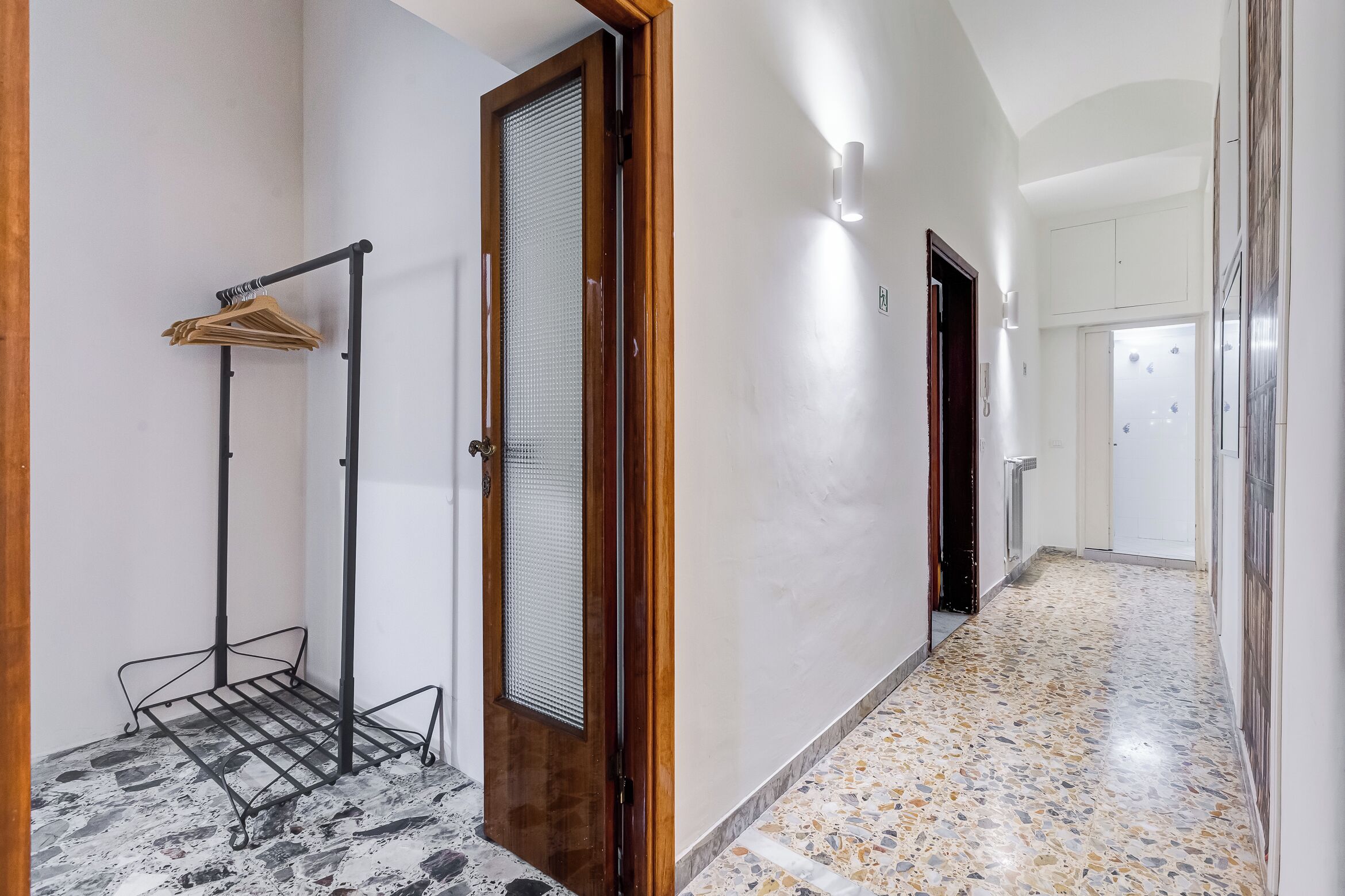 Un couloir avec un présentoir en métal, une porte en bois dotée d’un panneau de verre et un sol carrelé.
