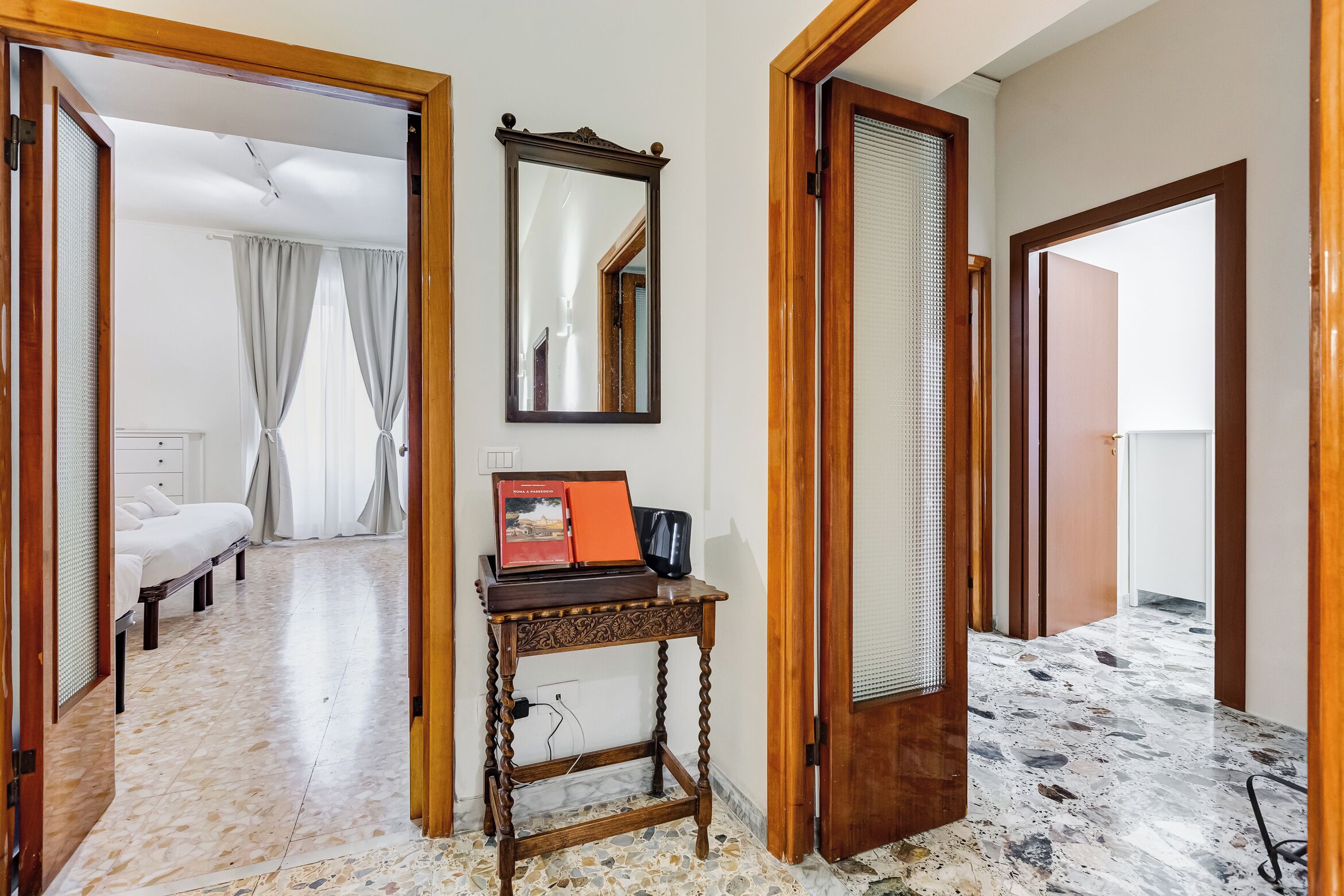 A hotel room with a bed, a bedside table with a mirror, a wooden door, and a tiled floor.