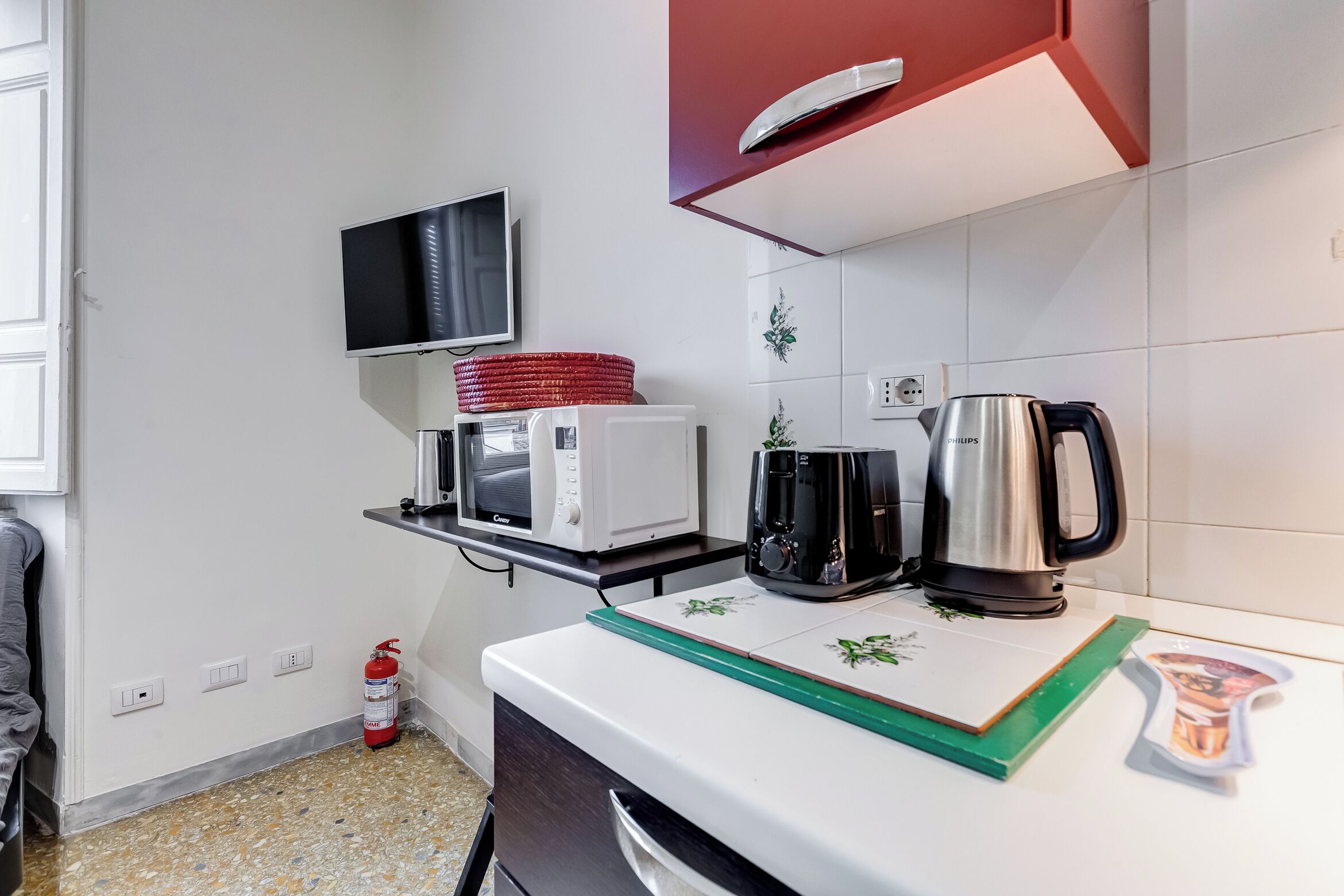 A compact kitchen with a microwave, coffee maker, and kettle on the counter, a fire extinguisher near the wall, and a television mounted above the microwave.