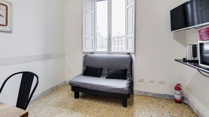 A small, compact room with a grey sofa, a television mounted on the wall, a fire extinguisher, and a window with a view of buildings.