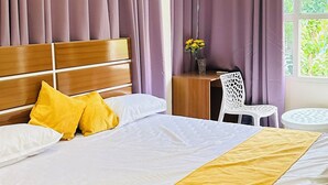 Un dormitorio con cama, cabecera de madera, almohadas amarillas, una mesita de noche con un jarrón de flores y cortinas moradas.