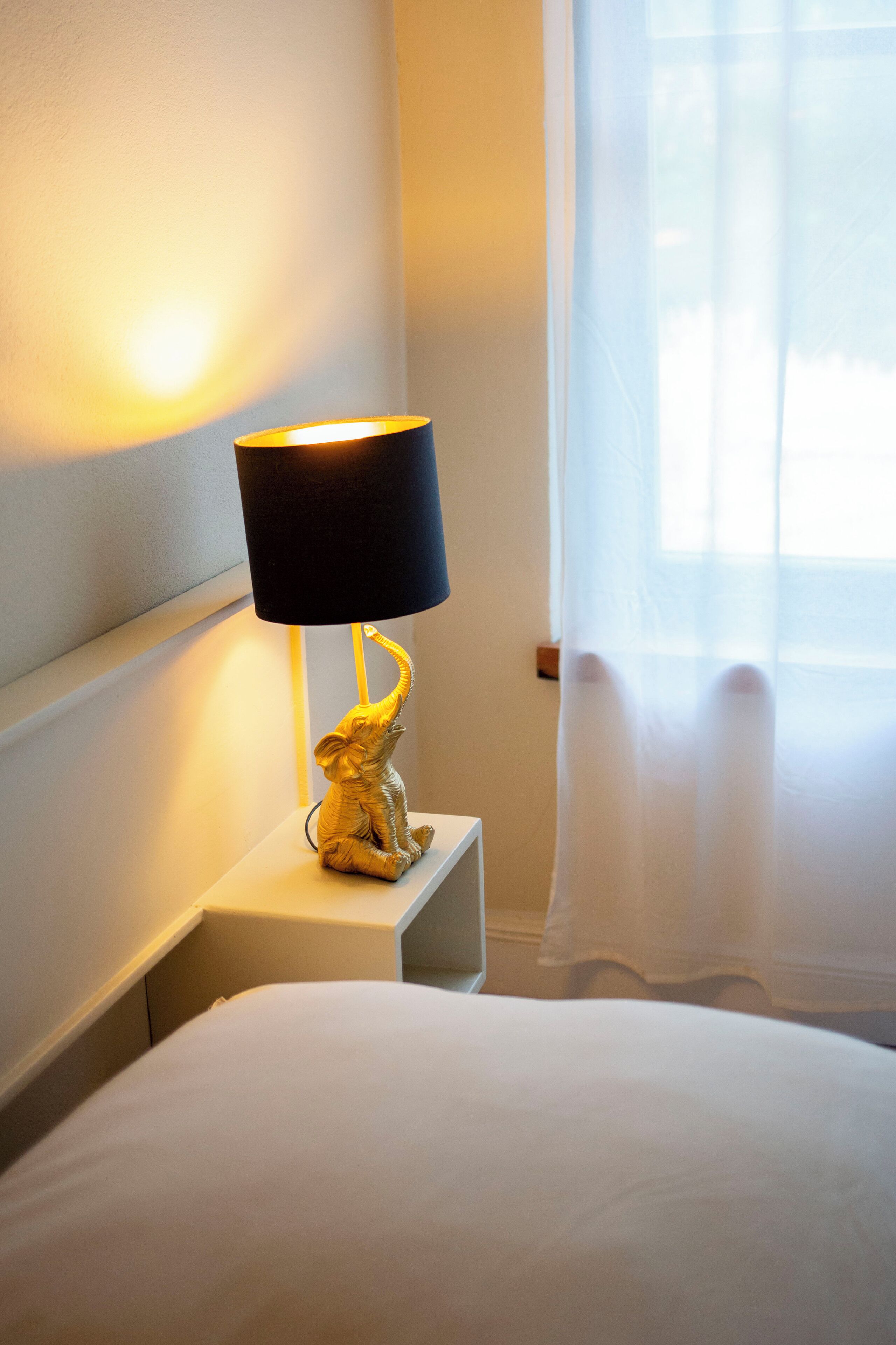 A bed with white bedding, a black lamp with a gold base, and a sheer curtain partially drawn to the side.